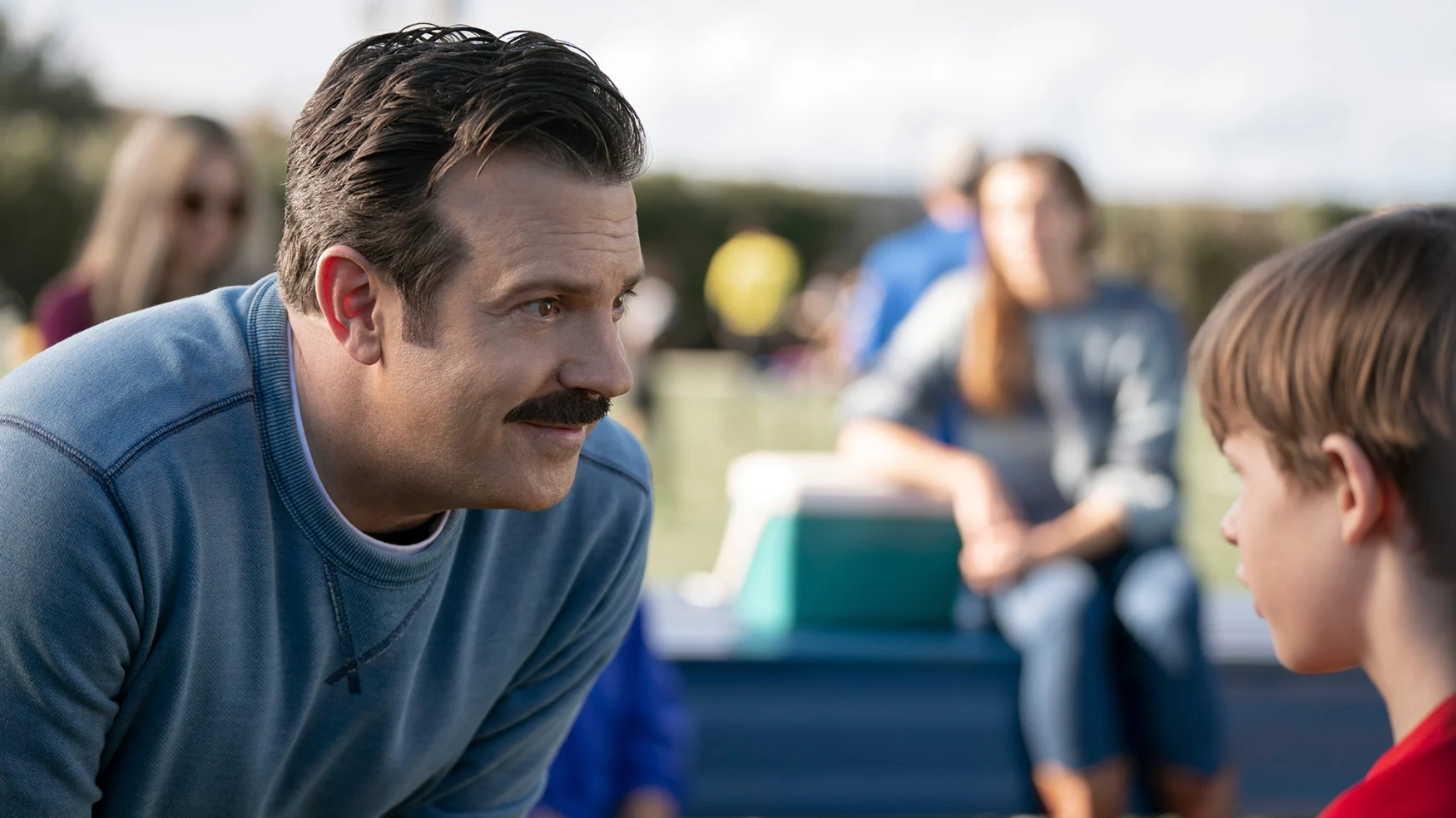 Jason Sudeikis as ‘Ted Lasso.’ He’s on a soccer field, leaning down and talking to a young boy.