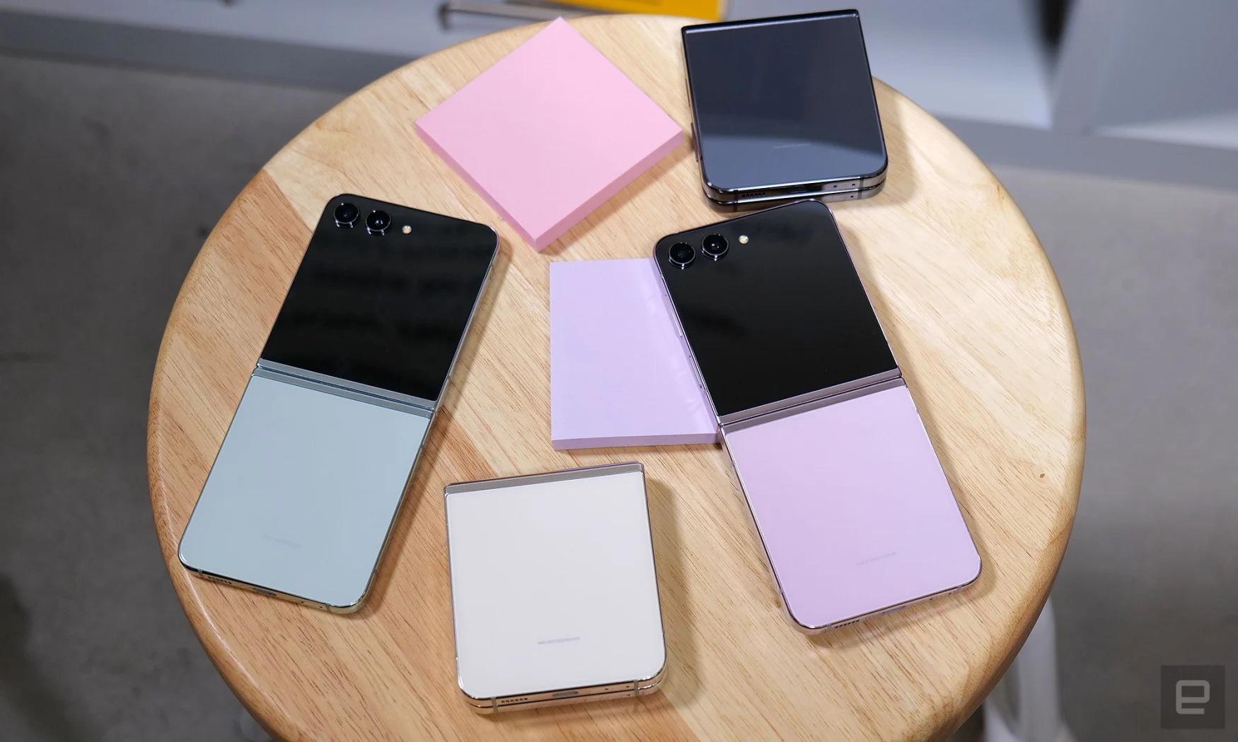 Four Samsung Galaxy Z Flip 5 units in graphite, lavender, cream and mint, laid out on a wooden stool. Two stick-it notepads are also on the surface.