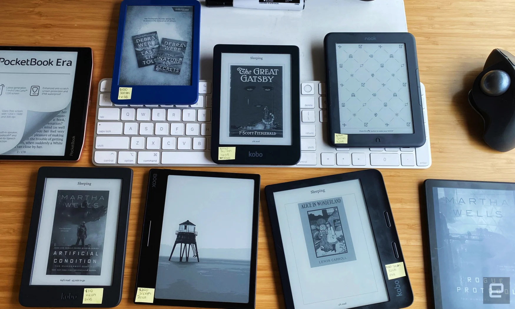 An assortment of ereaders, including ones from Kobo, Kindle, Boox, Nook and PocketBook are arranged on a tan bamboo desk with a white keyboard and black mouse in the background. 