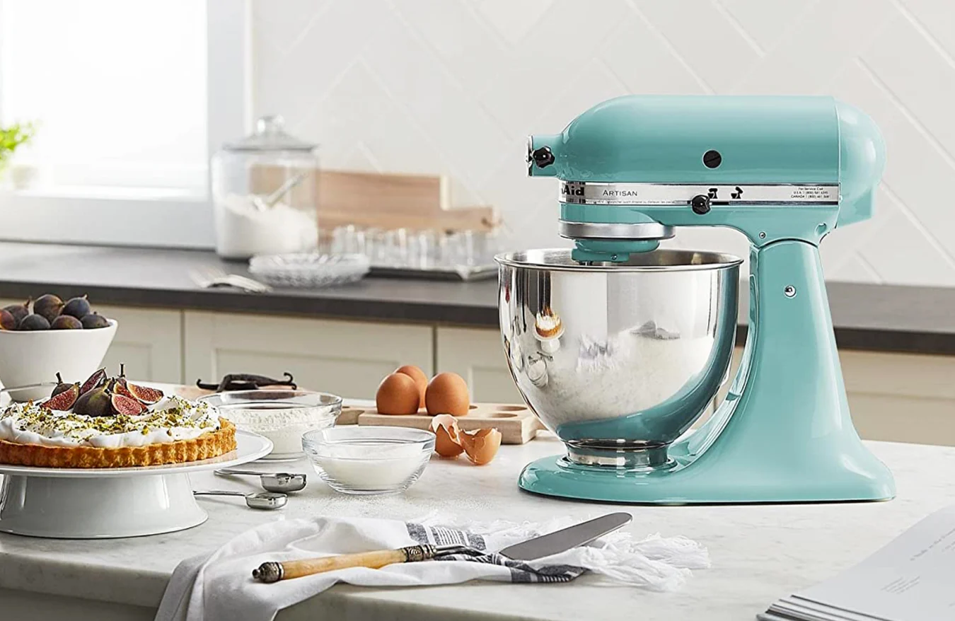 A KitchenAid Artisan series standing mixer on a countertop surrounded by baking supplies including eggs, a bowl of sugar, a spatula and a finished tarte.
