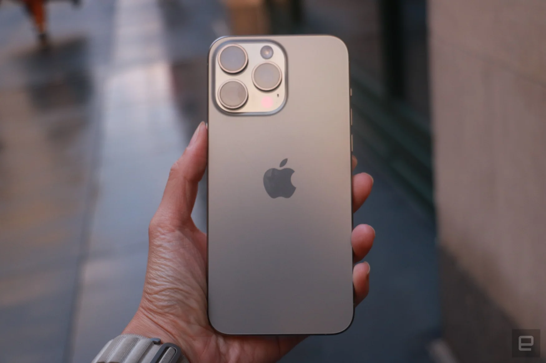 The iPhone 15 Pro Max held in mid-air with a sidewalk in the background.