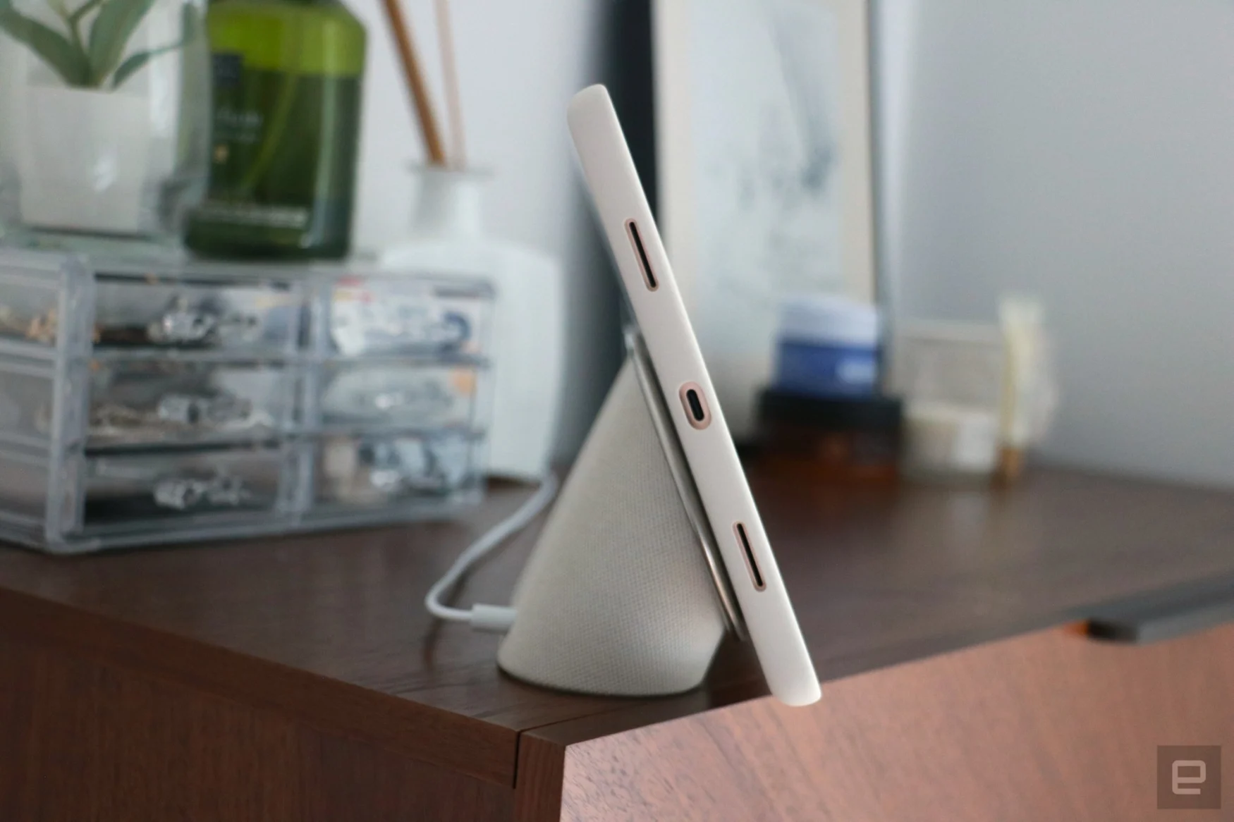 Tampilan samping Tablet Pixel di meja rias kayu kenari, menampilkan port USB-C di tepi kirinya.  Itu terpasang di dasar speaker, dengan casing pelindungnya menyala.
