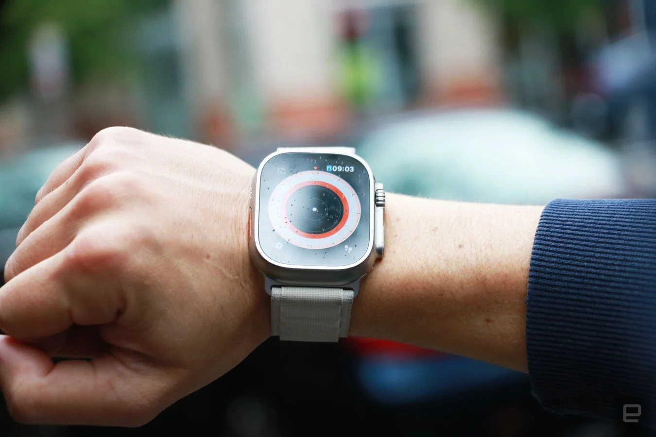Die Apple Watch Ultra mit alpinem Schlaufenband am Handgelenk einer Person.  Der Bildschirm zeigt die sogenannte technische Ansicht der Kompass-App mit einem weißen Punkt in der Mitte. 