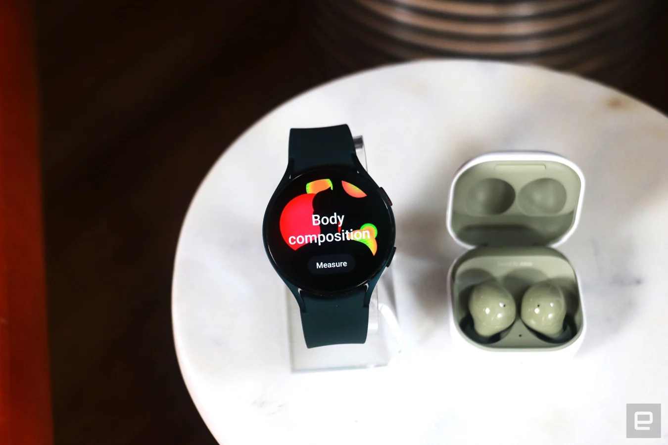 A black Galaxy Watch 4 on a stand next to a pair of olive Galaxy Buds 2. 