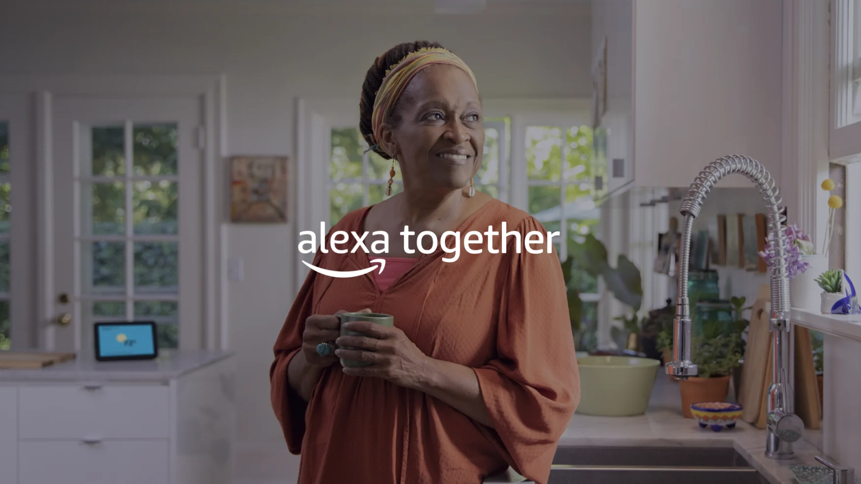 Eine Frau mit einem Becher, im Hintergrund ein Alexa-Display auf einem Küchentisch.  Die Wörter 