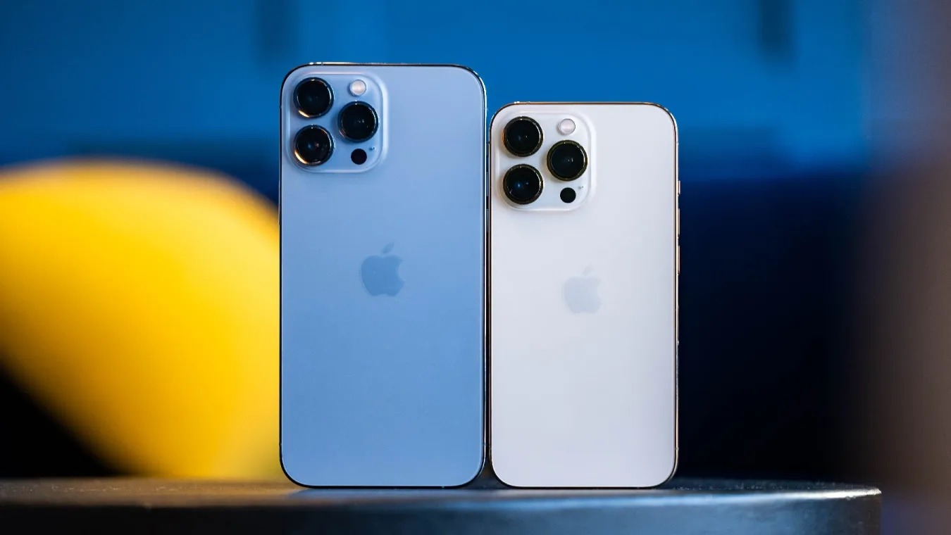 Ein blaues iPhone 13 Pro Mac und ein goldenes iPhone 13 Pro nebeneinander auf einem Tisch, die Rückkameras sind uns zugewandt.