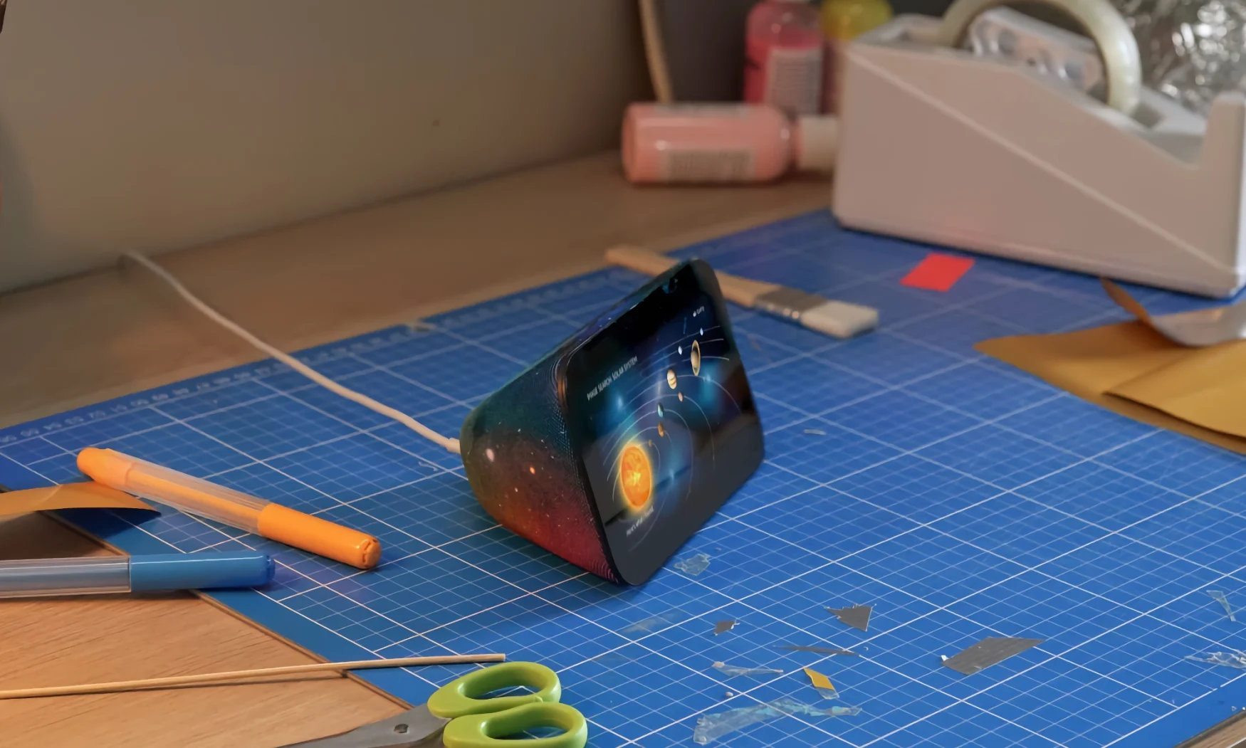 Amazon marketing photo of the Amazon Echo Show 5 Kids with a galaxy design on its back and an illustration of the solar system on its screen. It sits on a blue drafting mat on a desk with scissors, pens and other supplies nearby.