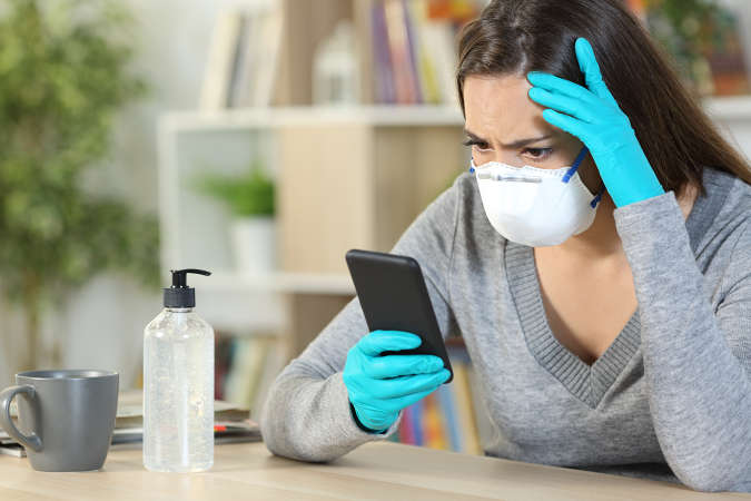 Worried girl with latex gloves and protective mask reads covid-19 news on smart phone sitting at home