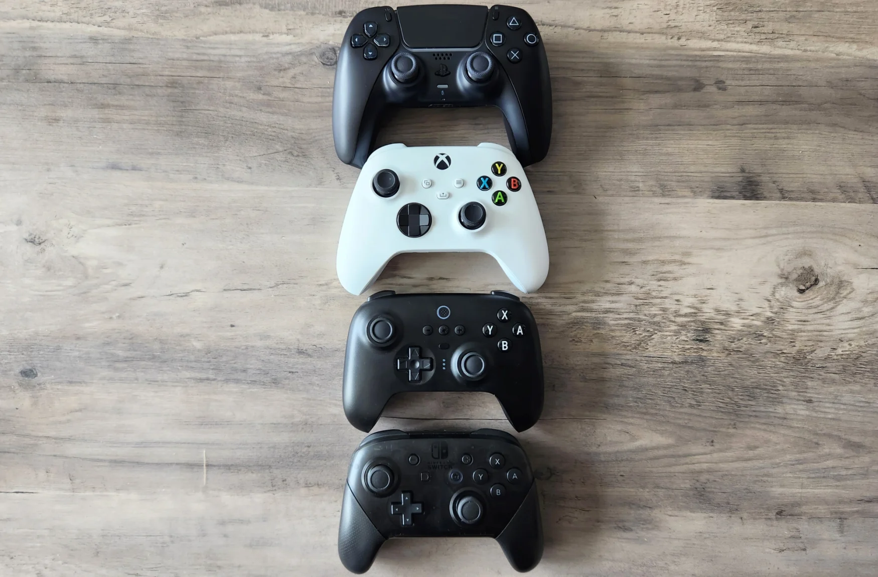 Four video game controllers rest on a wooden tabletop, one above the other. From the top: the Sony DualSense PlayStation 5 Controller, the Microsoft Xbox Series X/S Controller, the 8BitDo Ultimate Bluetooth Controller and the Nintendo Switch Pro Controller.