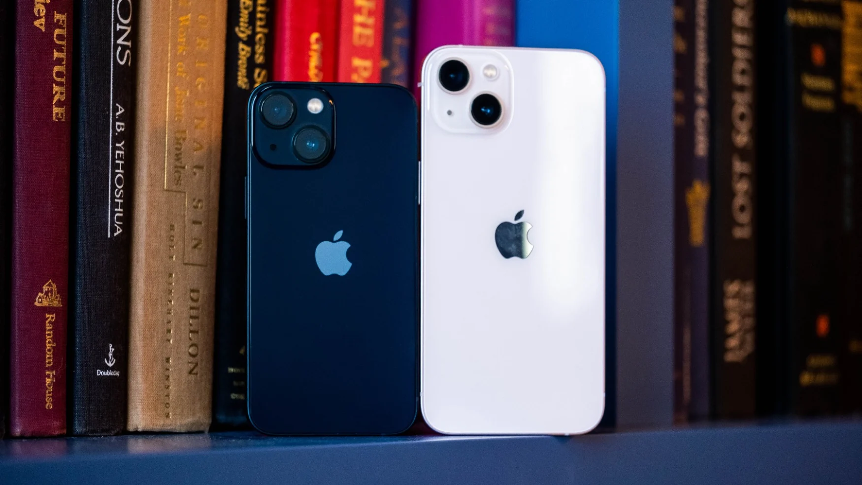 A black iPhone 13 mini and pink iPhone 13 standing in front of some books on a shelf, with their rears facing the camera.