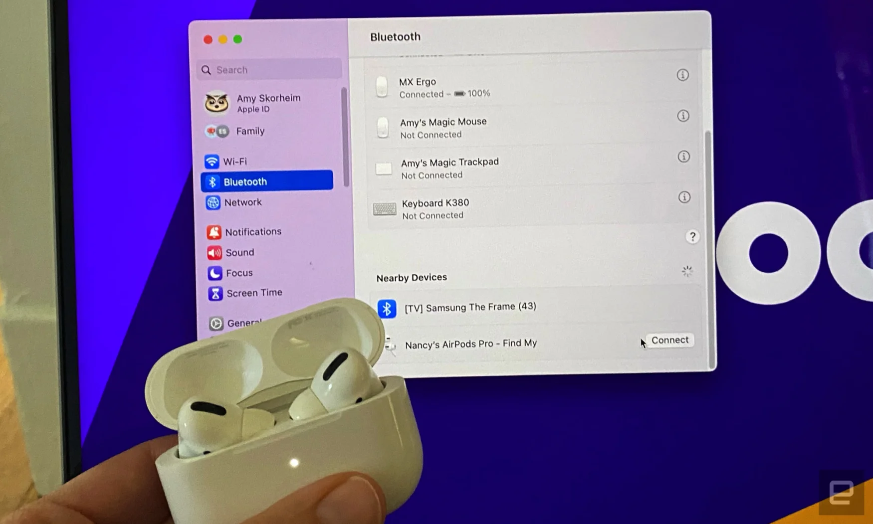 El estuche de los AirPods con la luz de emparejamiento blanca brillante se coloca cerca de la pantalla del MacBook con el menú de Bluetooth abierto. 