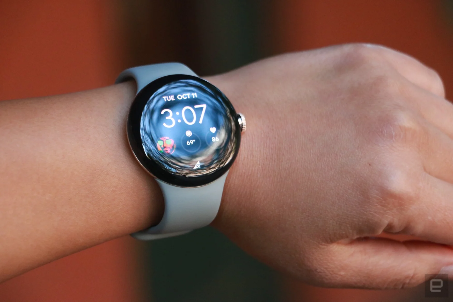 The Pixel Watch on a wrist held up against a brick red background. On its screen is the time and three complications below it.