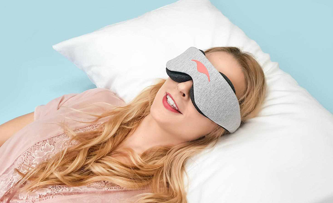 A smiling woman is laying down in bed while wearing a Manta Sleep Mask.