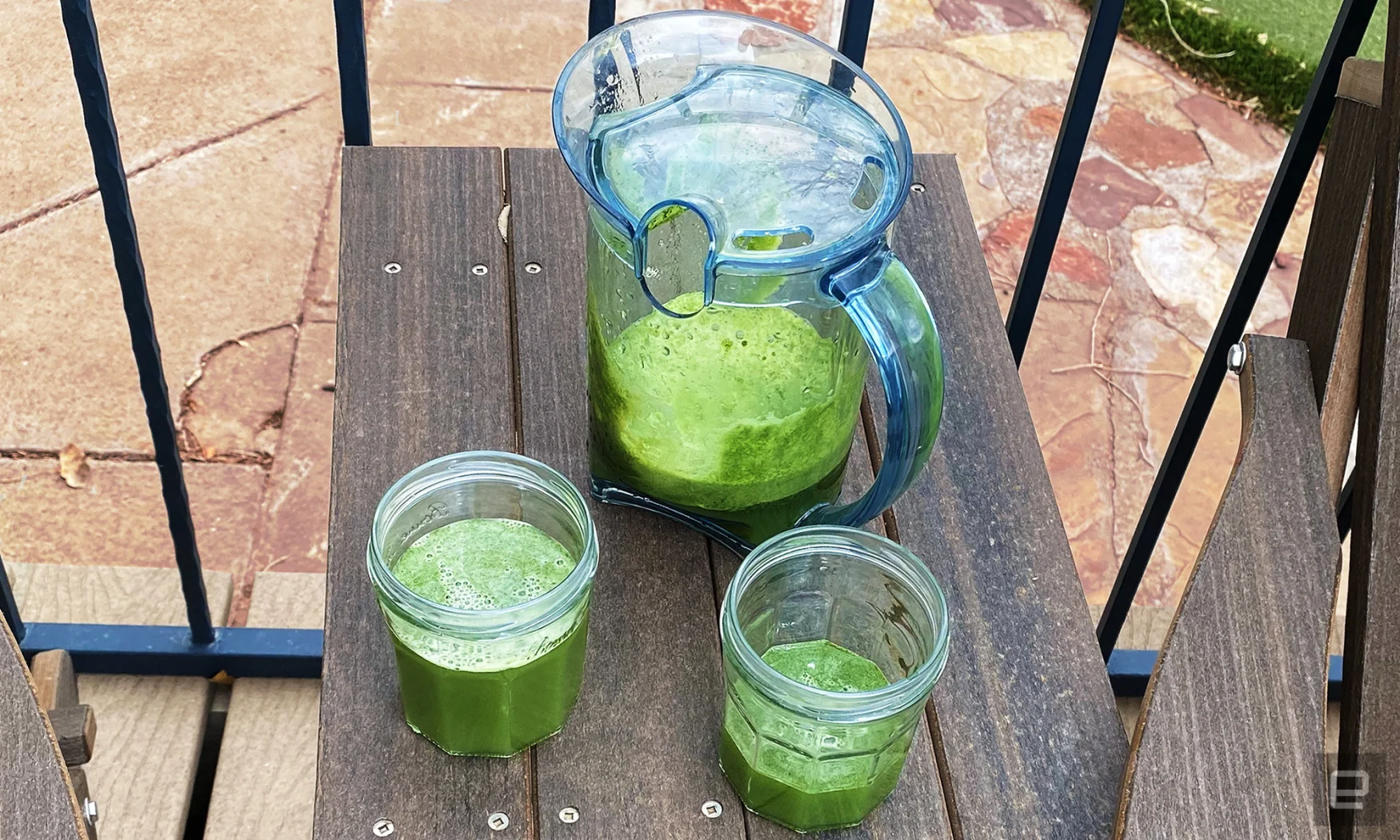 A pitcher and two mugs sit on a brown wooden table outside.  They are filled with green juice made from kale, apples, and celery. 