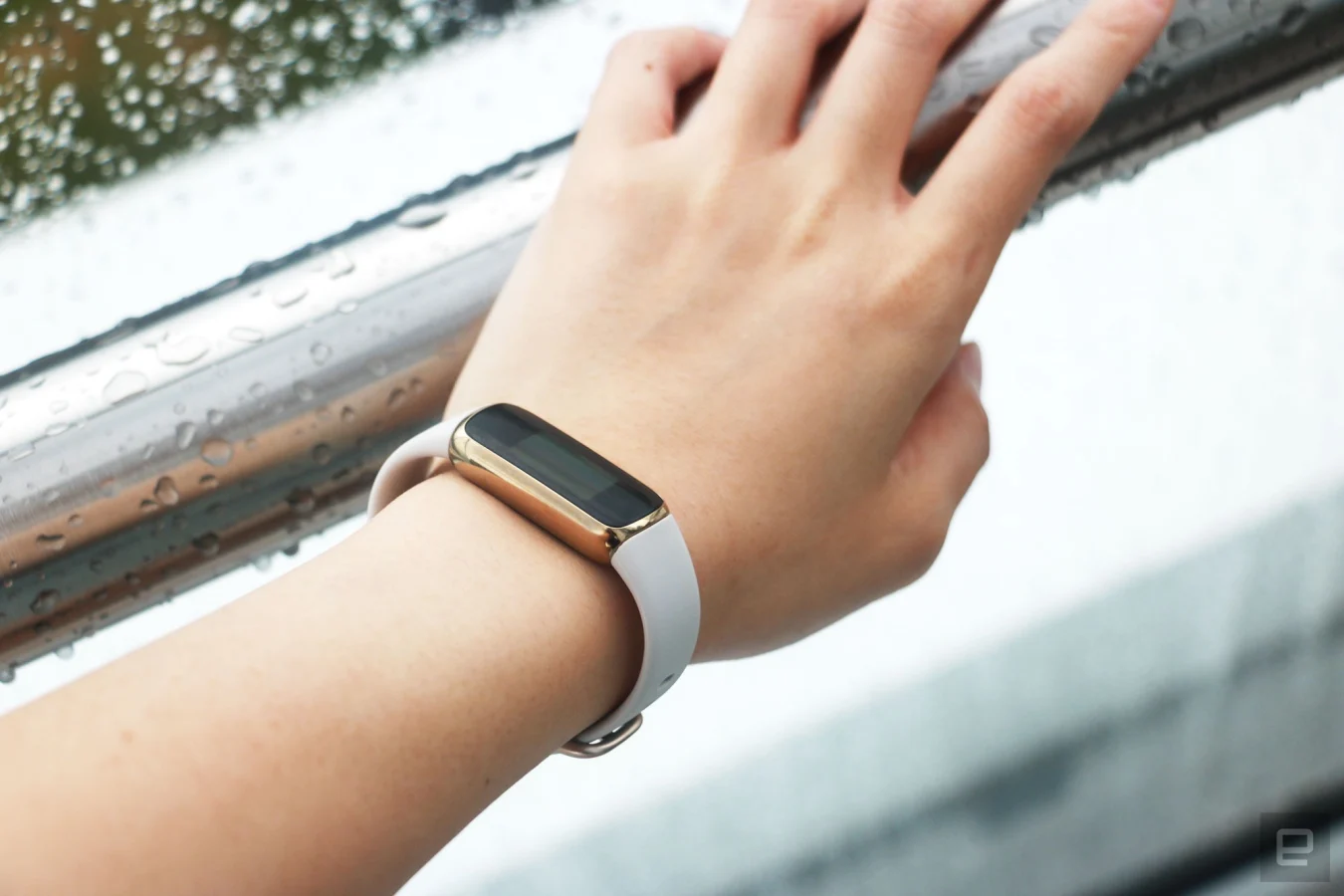 The Fitbit Luxe with a light pink silicone band on a wrist leaning on a wet railing. The screen is off.
