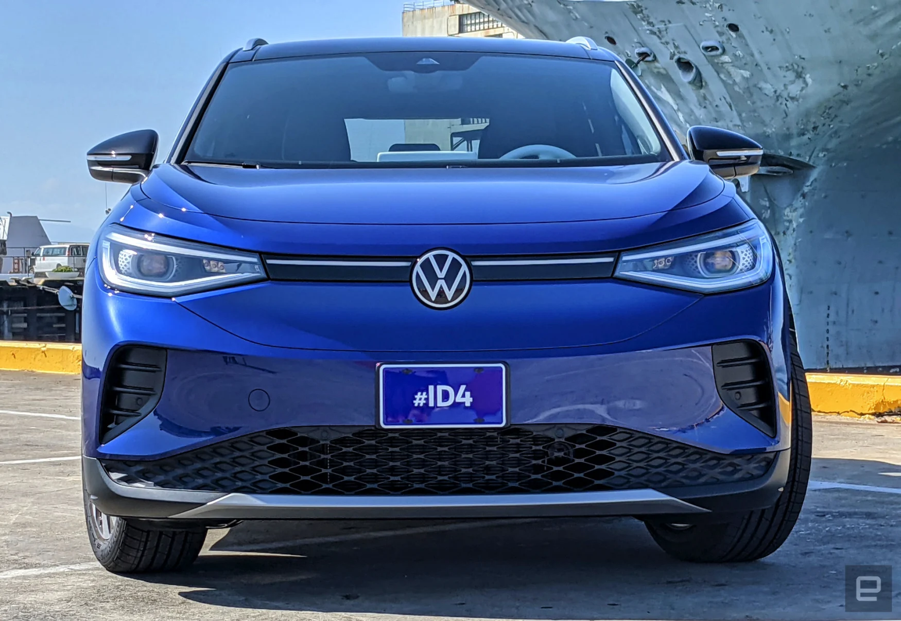 A blue Volkswagen ID.4 EV is parked in a lot right next to San Francisco docks with military ships.