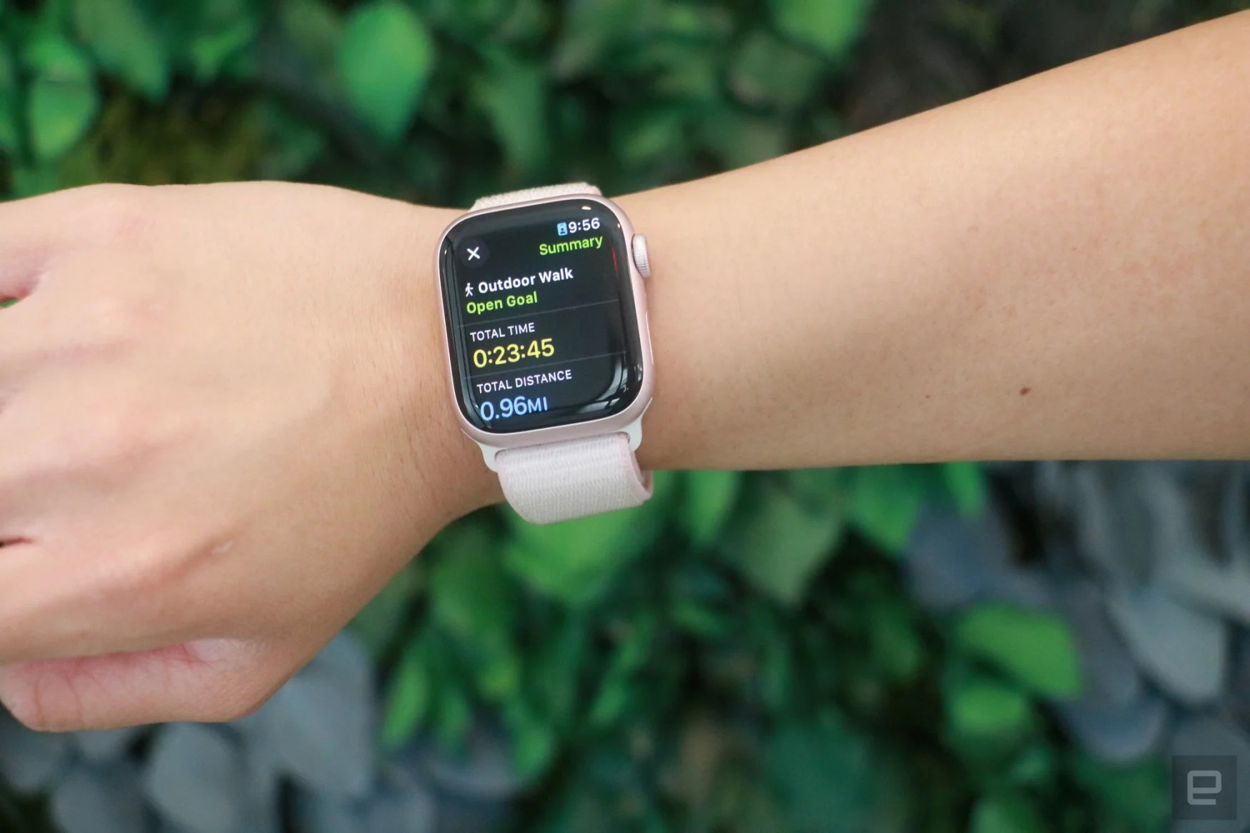 Apple Watch Series 9, yeşil yeşilliklerin önünde bir kişinin bileğinde, açık havada yürüyüş antrenmanının özetini gösteriyor.