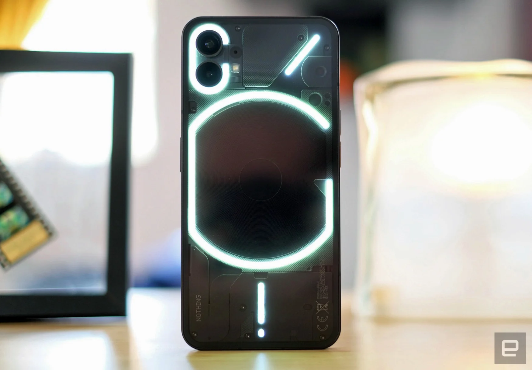 The new Nothing Phone 1 stands on its end on a table. The phone is black with a see-through back showing a glowing series of lines and rings.
