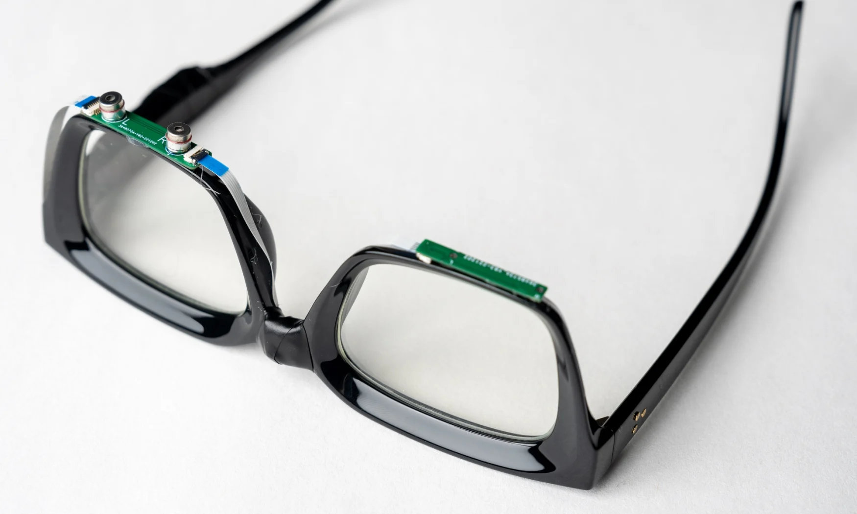 A pair of black-plastic-framed eyeglasses sitting upside down on a white surface. The frames have small circuit boards with tiny speakers and microphones attached below the lenses.
