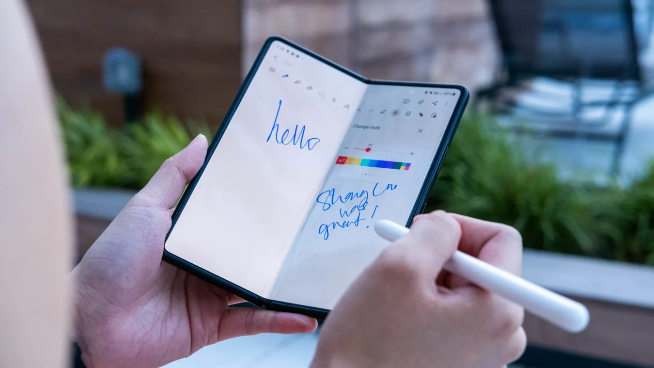 A person using the S Pen Pro to draw on the Samsung Galaxy Z Fold 3. They are holding the phone, half-folded, in their left hand with an S Pen Pro in their right. On the screen, they've scrawled 