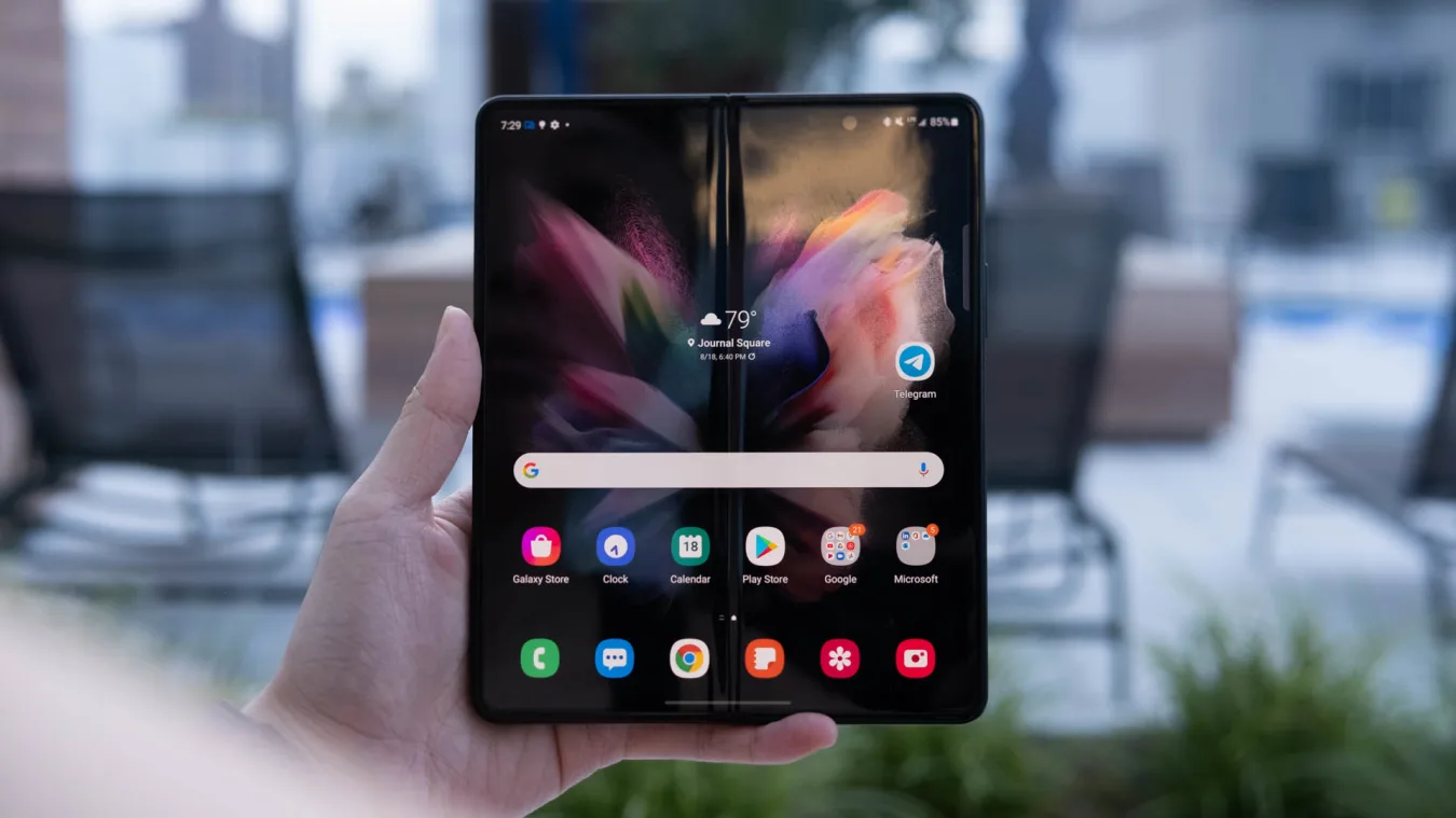 A hand holding up the Samsung Galaxy Z Fold 3 with its screen open and facing the camera.  A crease runs down the middle of the home page. 