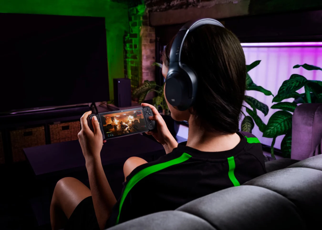 A person wearing headphones and sitting on a couch while holding a Razer Edge handheld gaming device.