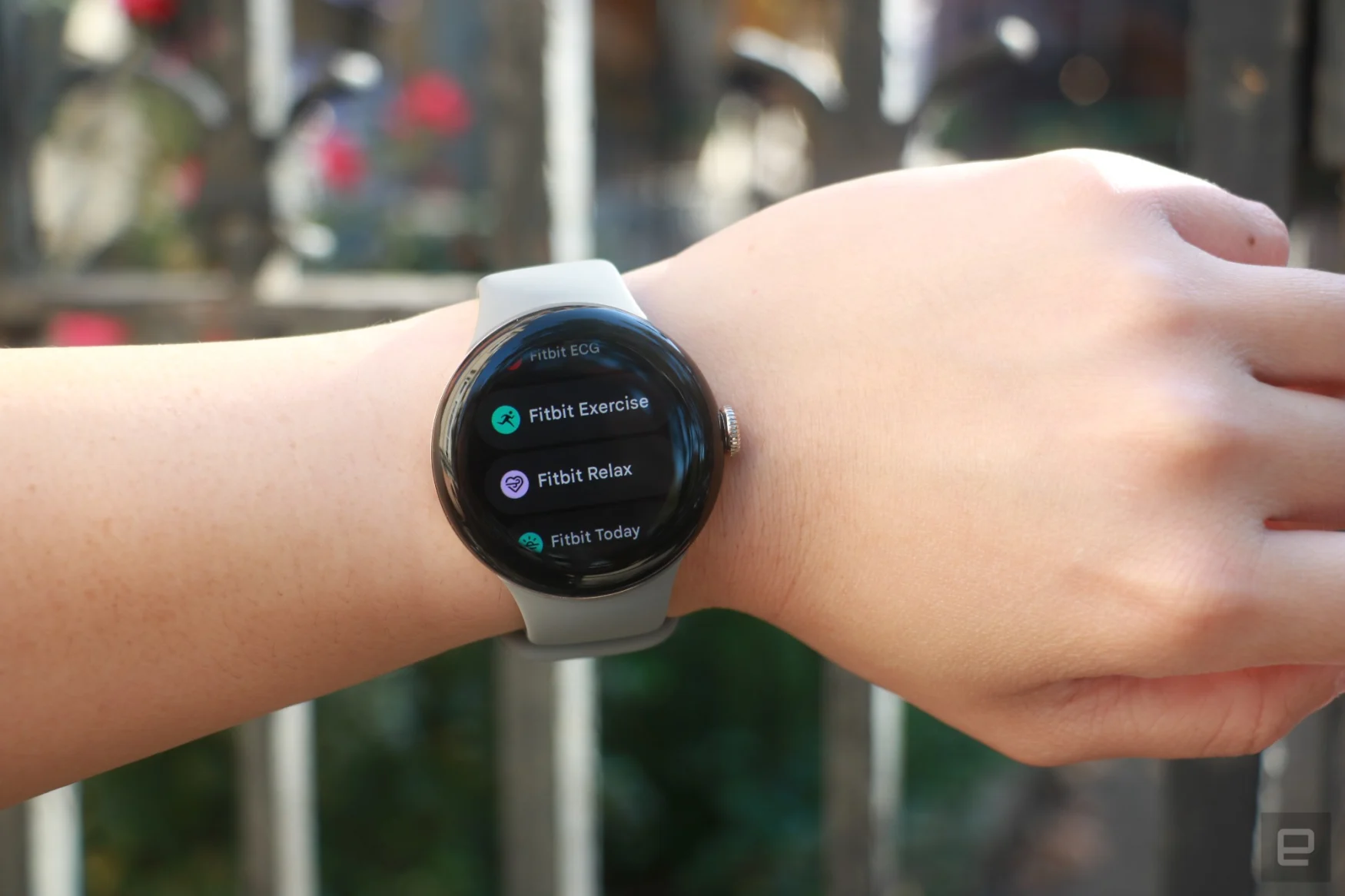 The Pixel Watch 2 on a person's wrist, showing a list of Fitbit apps. From top to bottom, they are Fitbit ECG, Fitbit Exercise, Fitbit Relax and Fitbit Today.