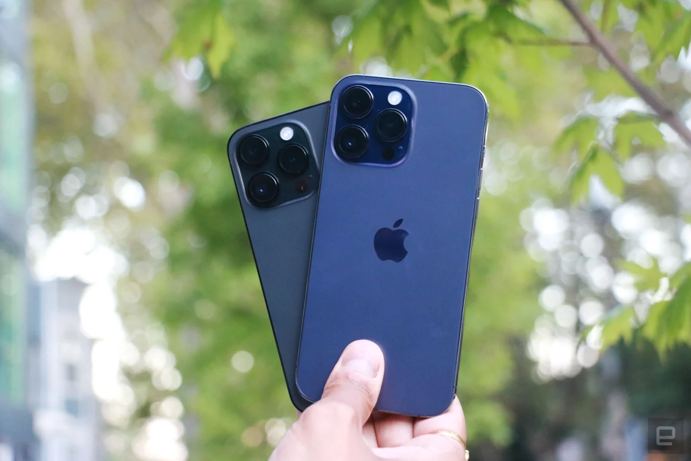 The purple iPhone 14 Pro Max and the black iPhone 14 Pro held on top of each other in one hand, slightly fanned out so you can see the triple rear cameras of each handset.
