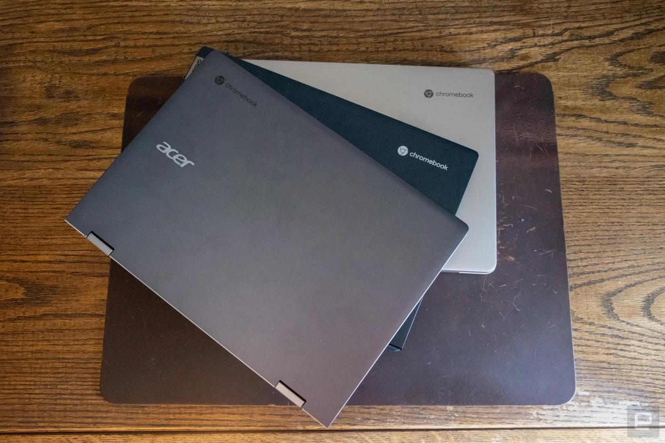 Three Chromebook laptops stacked on top of each other, fanning out a bit, on top of a desk protector on a wooden table.