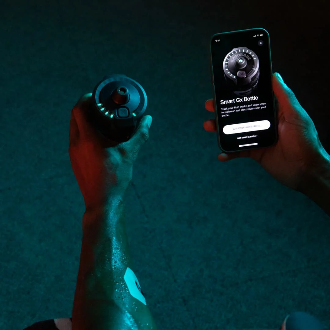 A person holds the Gatorade Smart Gx Bottle, which has a lid with lights around the rim. In the other hand they hold a smartphone showing the Gx app with the text “Smart Gx Bottle”.