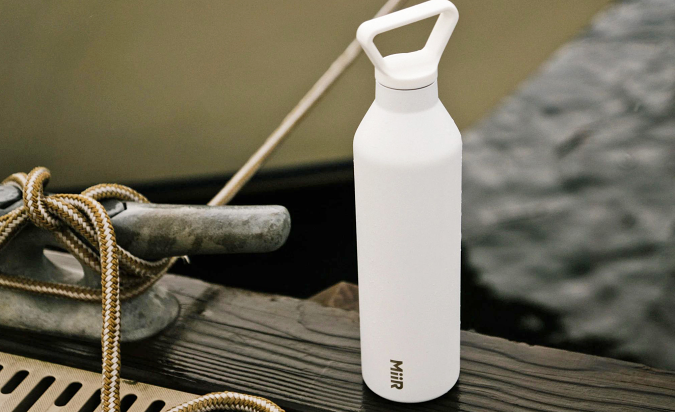 A white MiiR Insulated bottle sits on the edge of a dock with the water and a boat in the background.
