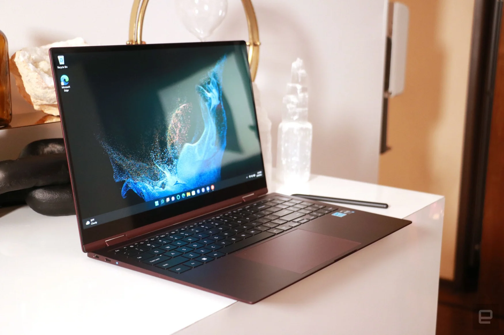 A burgundy Samsung Galaxy Book 2 Pro 360 on a white desk with decorative ornaments behind it.