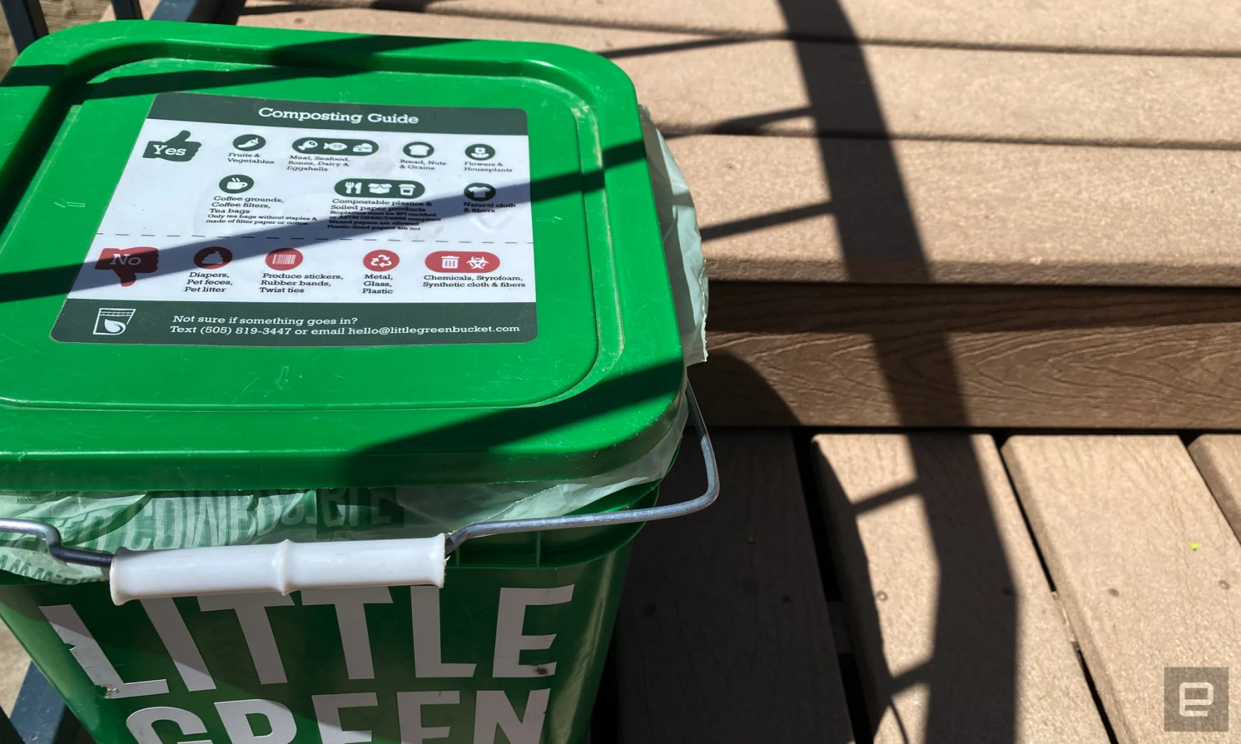 A green, square compost bucket sits on wooden steps. The instructions for what foods can go in the bucket are listed on the lid. 