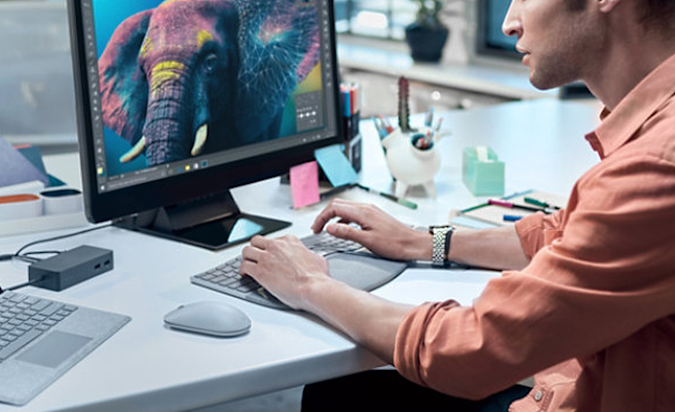 A man sitting at a desk in front of a computer with his hands on the Microsoft Surface Ergonomic Keyboard.