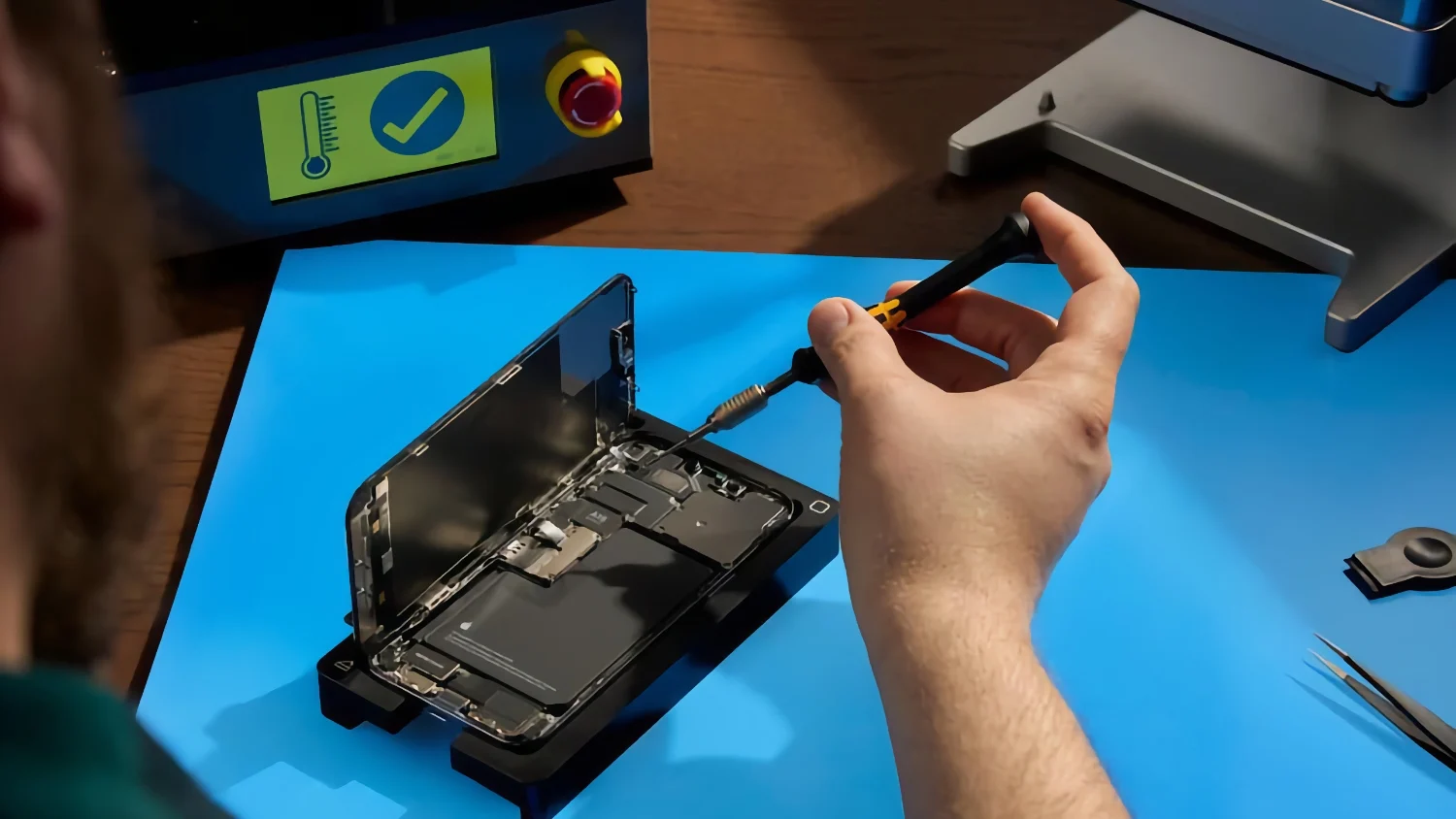 A person’s hand holding a tool, repairing an opened-up iPhone on a blue mat.