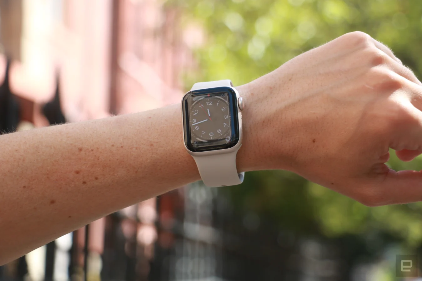 The Apple Watch SE (2022) on a person's wrist held up in front of a red building and a tree.