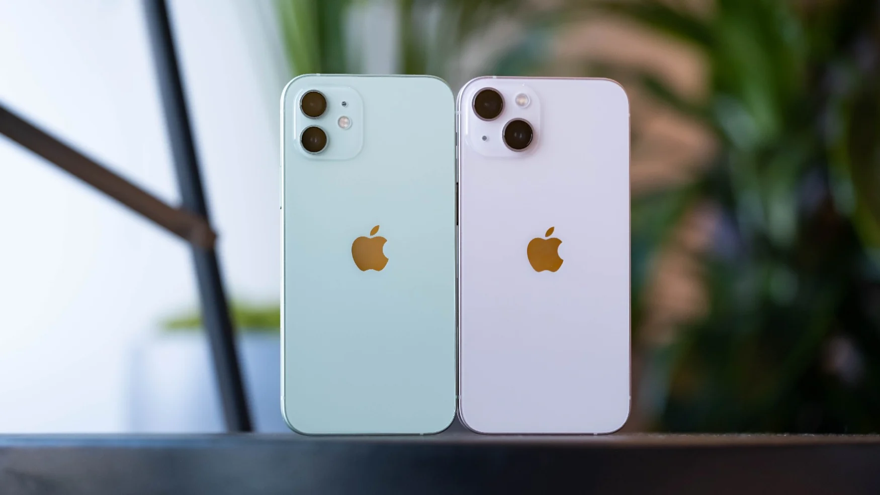 A green iPhone 12 and pink iPhone 13 with their backs facing the camera. You can see their dual cameras. On the left, the pair of sensors are stacked on top of each other. On the right, they're laid out diagonally. The module on the right is also slightly wider and taller, and the cameras themselves are slightly bigger.