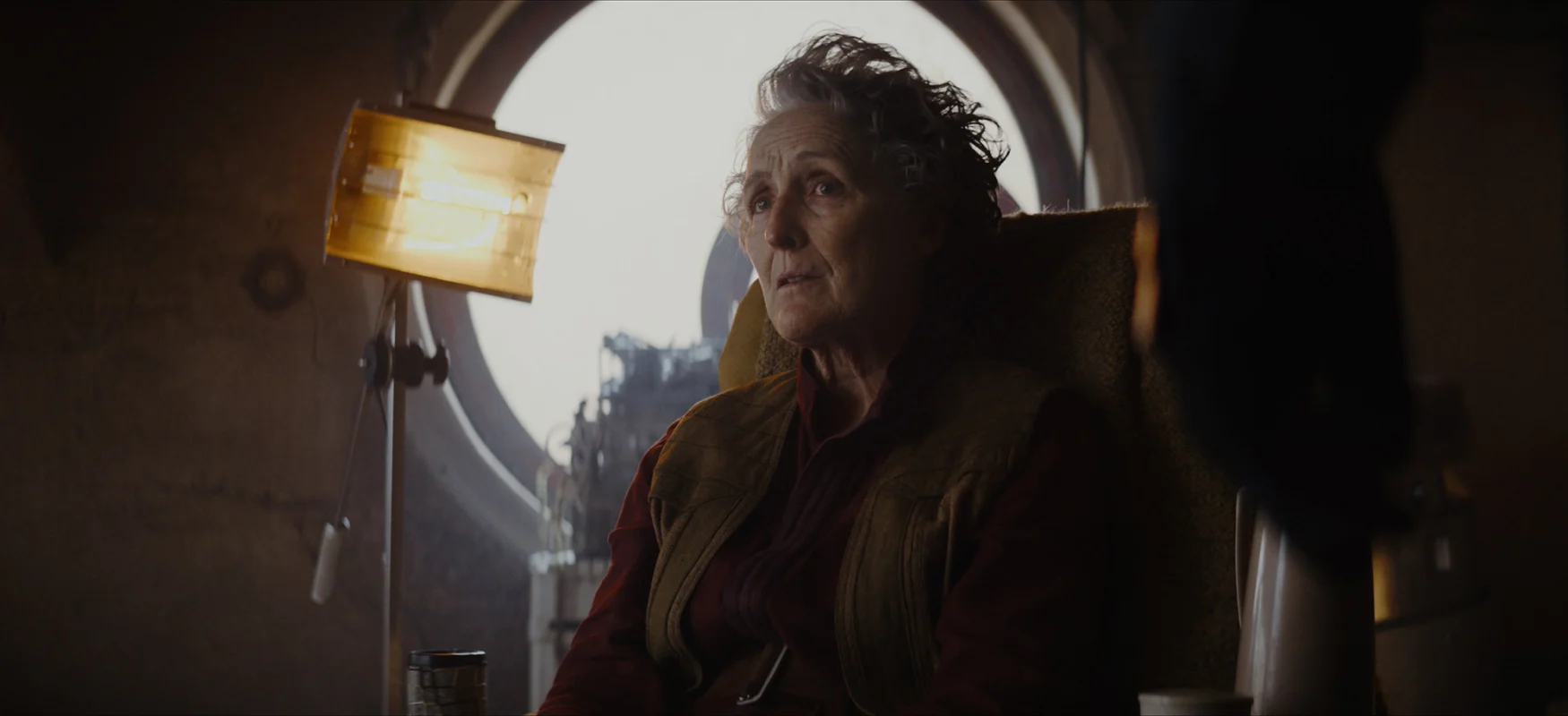 Maarva (Fiona Shaw) in ‘Andor.’ She sits in a fairly dark room with crude lighting equipment and a bright window behind her.