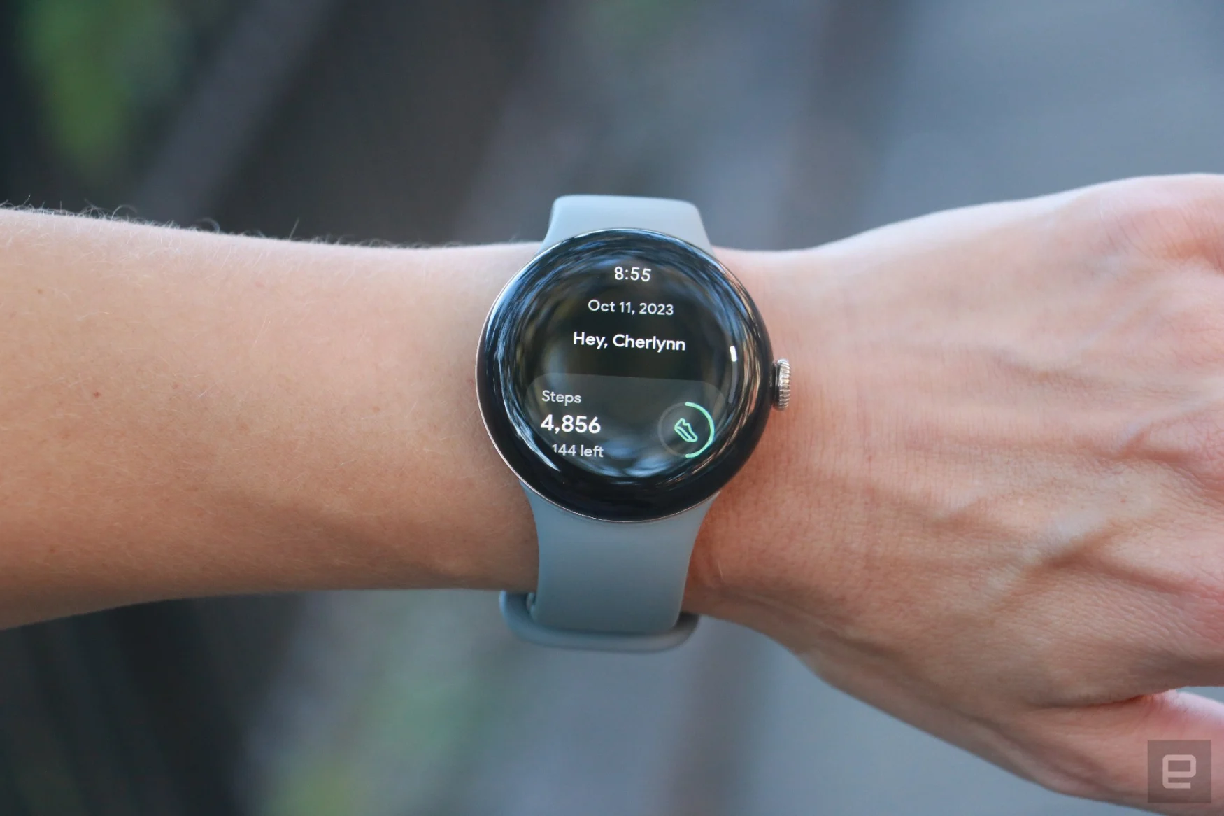 The Pixel Watch 2 on a person's wrist with a sidewalk and some grass in the background. The screen shows the Fitbit Today dashboard with the words 