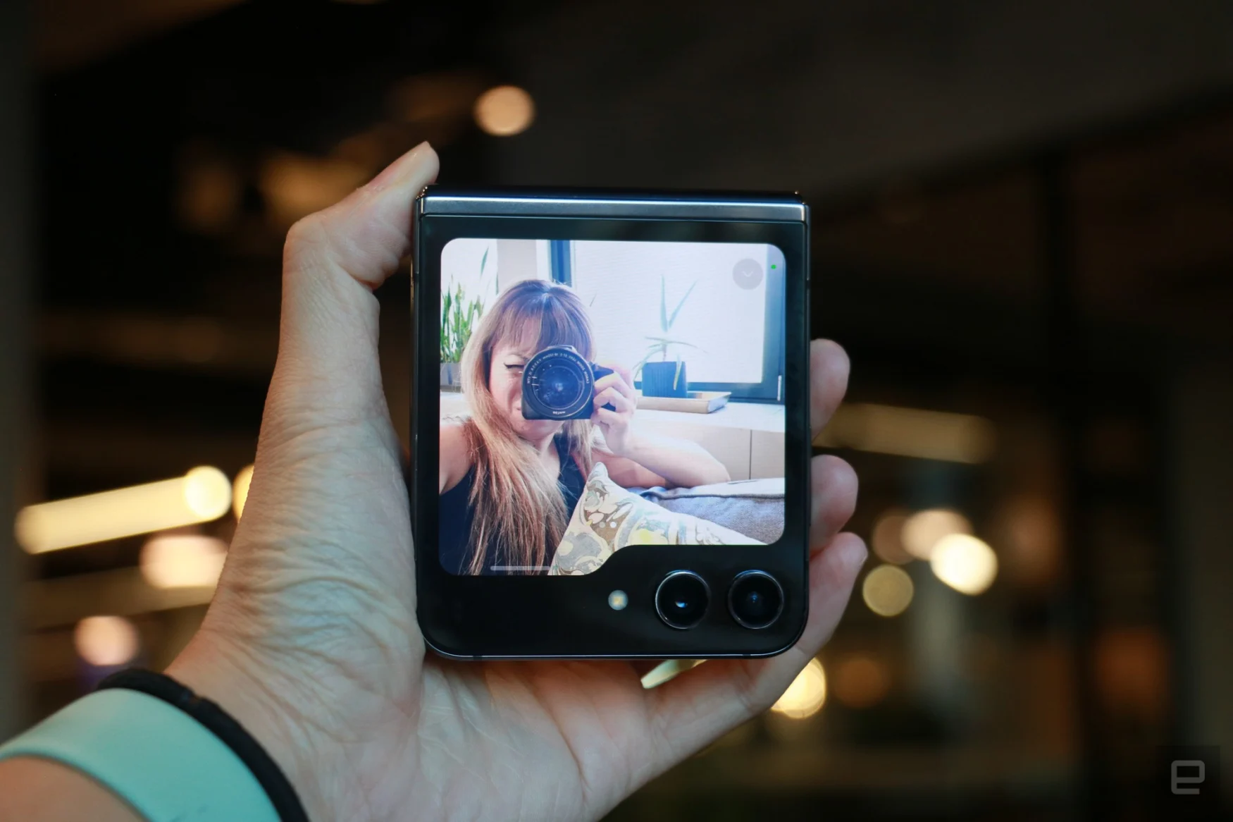 The Samsung Galaxy Z Flip 5 folded shut, held in mid-air. Its external screen shots a person taking a photo of the camera viewfinder feature.
