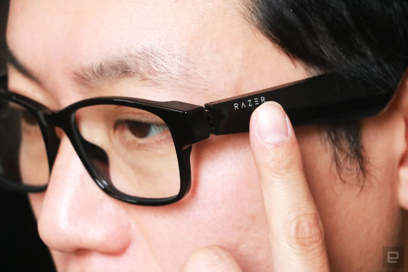 Razer Anzu smart glasses review photo. Close up of the left side profile of a man wearing the Razer smart glasses with his left index finger touching the left arm right below the Razer logo. 