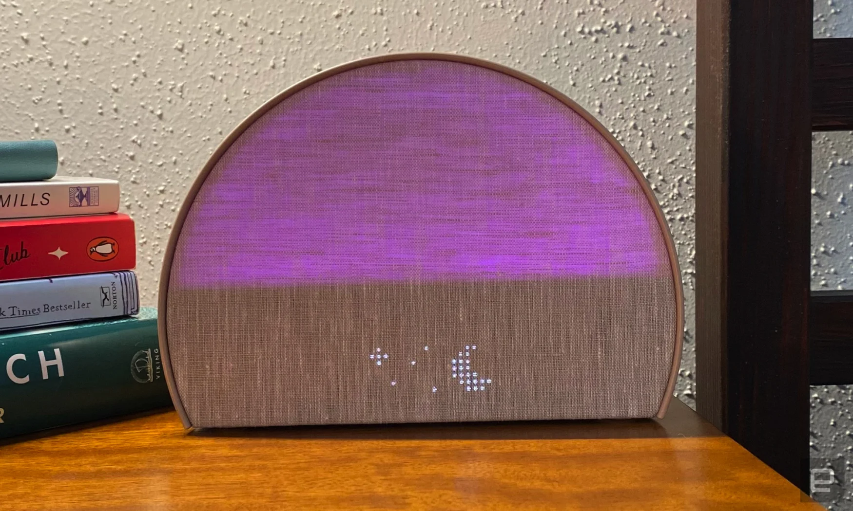 The Hatch Restore 2 lights up purple and displays a dot-matrix LED moon and stars. The unit sits on a wooden bedside table and the corner of a few books are stacked in the background. 