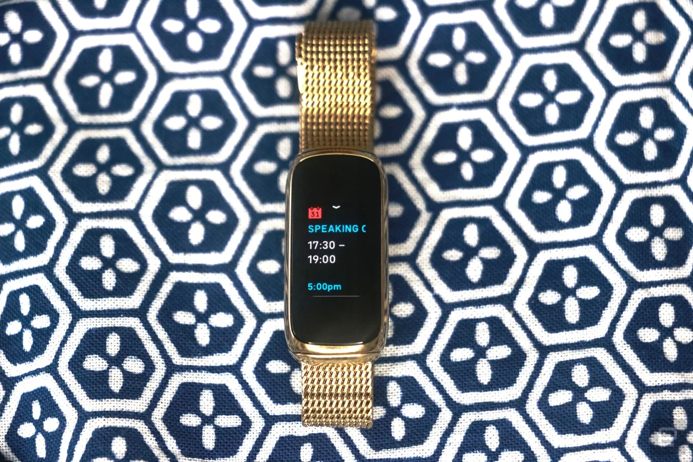 Front view of the Fitbit Luxe with a gold mesh bracelet on a patterned blue and white background. Its screen shows a calendar notification for an event from 5:30pm to 7pm.