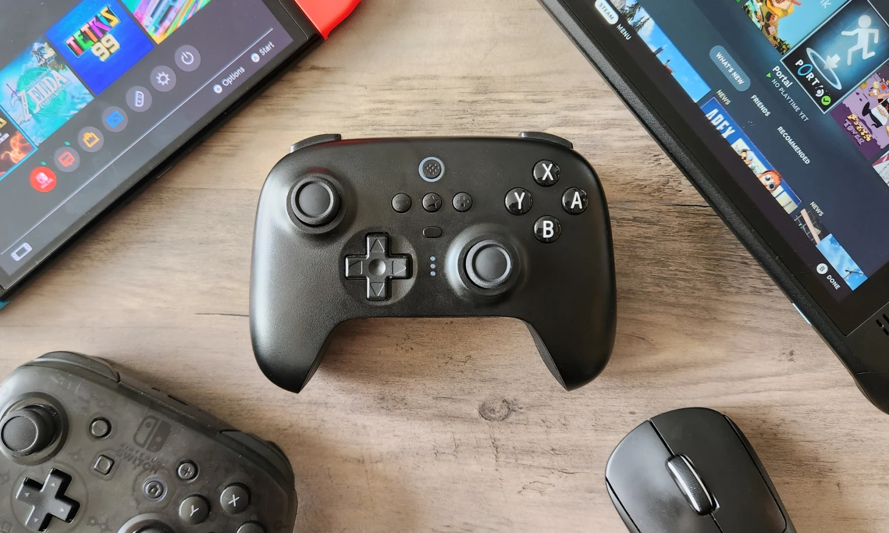 A black video game controller, 8BitDo Ultimate Bluetooth Controller, rests on a wooden table alongside a Nintendo Switch, a Nintendo Switch Pro Controller, a Valve Steam Deck, and a Logitech gaming mouse.
