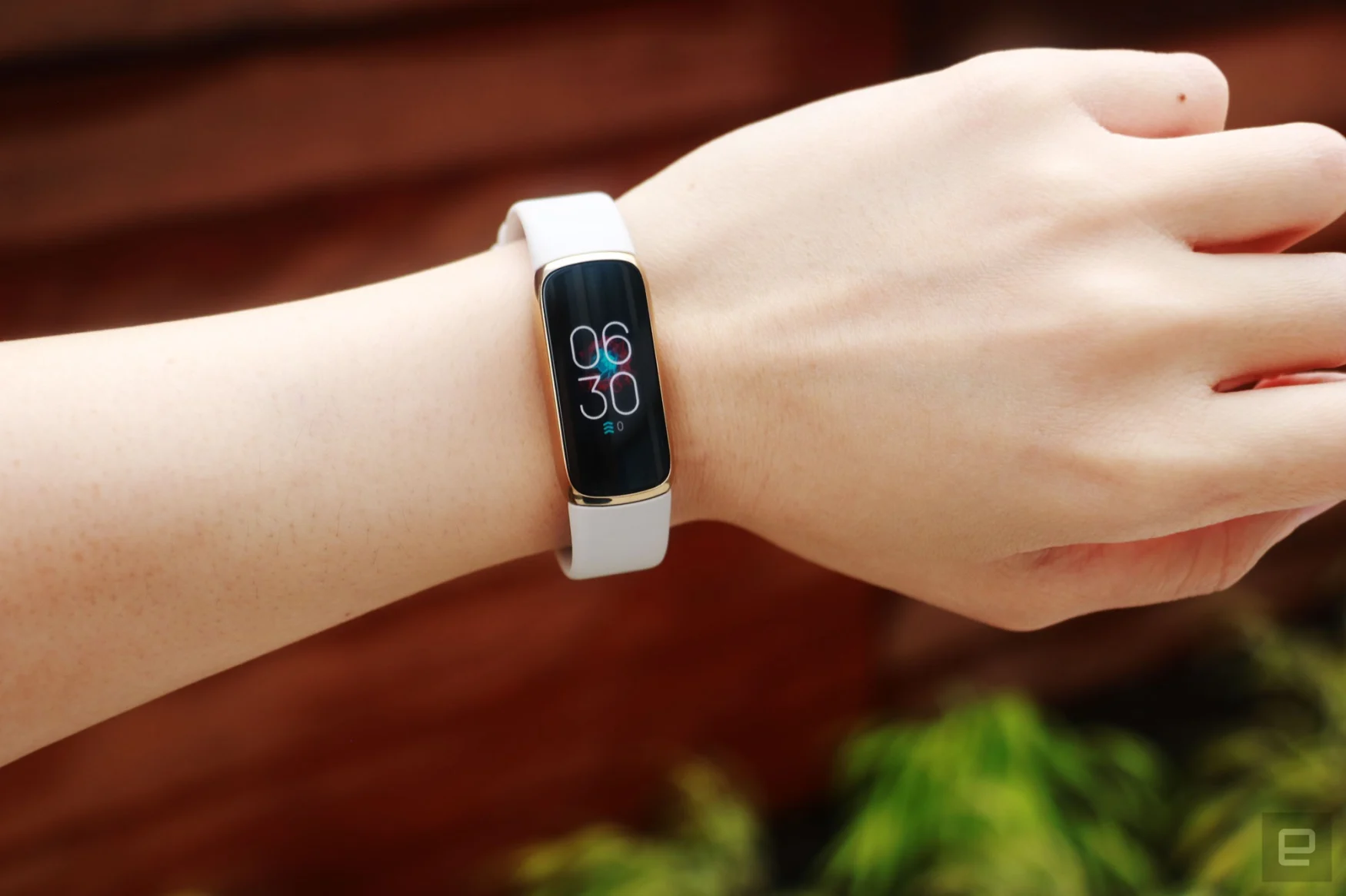 Front view of the Fitbit Luxe with a light pink silicone band on a wrist against a dark brown background with some greenery. The screen shows the time is 6:30pm.
