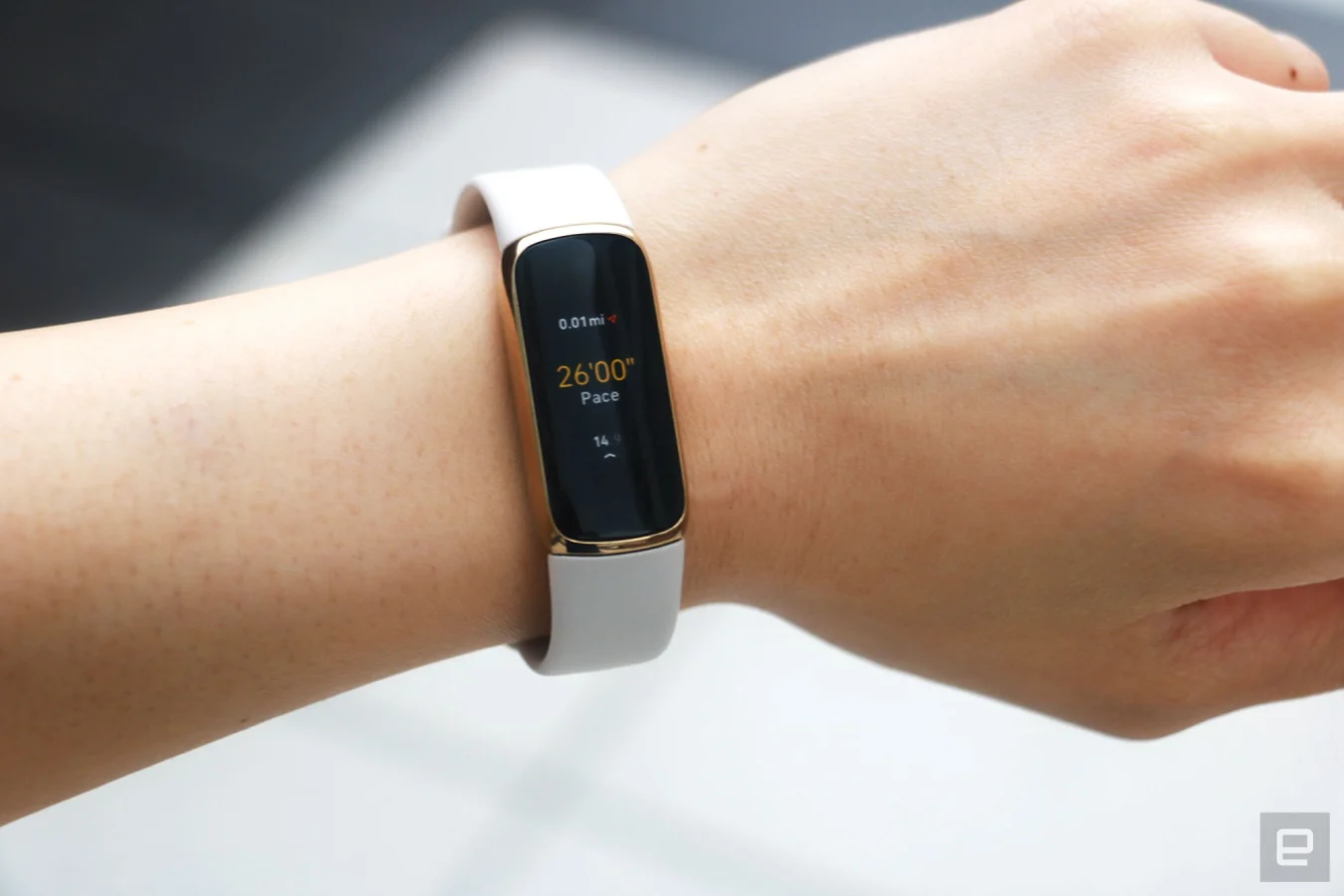 The Fitbit Luxe with a light pink silicone band on a wrist against a concrete gray background. The screen shows a run being tracked with a pace of 26:00 and 0.01 miles traveled.