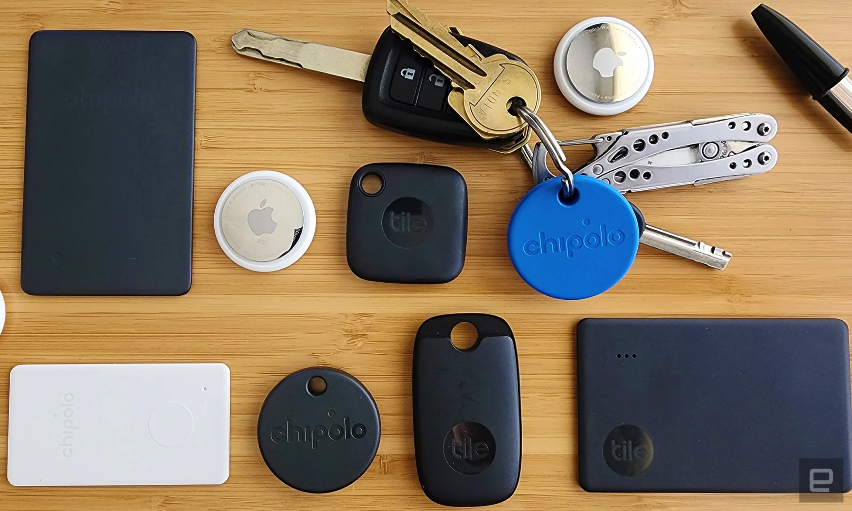 An assortment of bluetooth trackers arranged in a grid on a wooden background. Trackers include black Tile trackers in various shapes, two silver and white AirTag trackers and a round blue Chipolo tracker attached to a set of keys with a multitool key chain. 