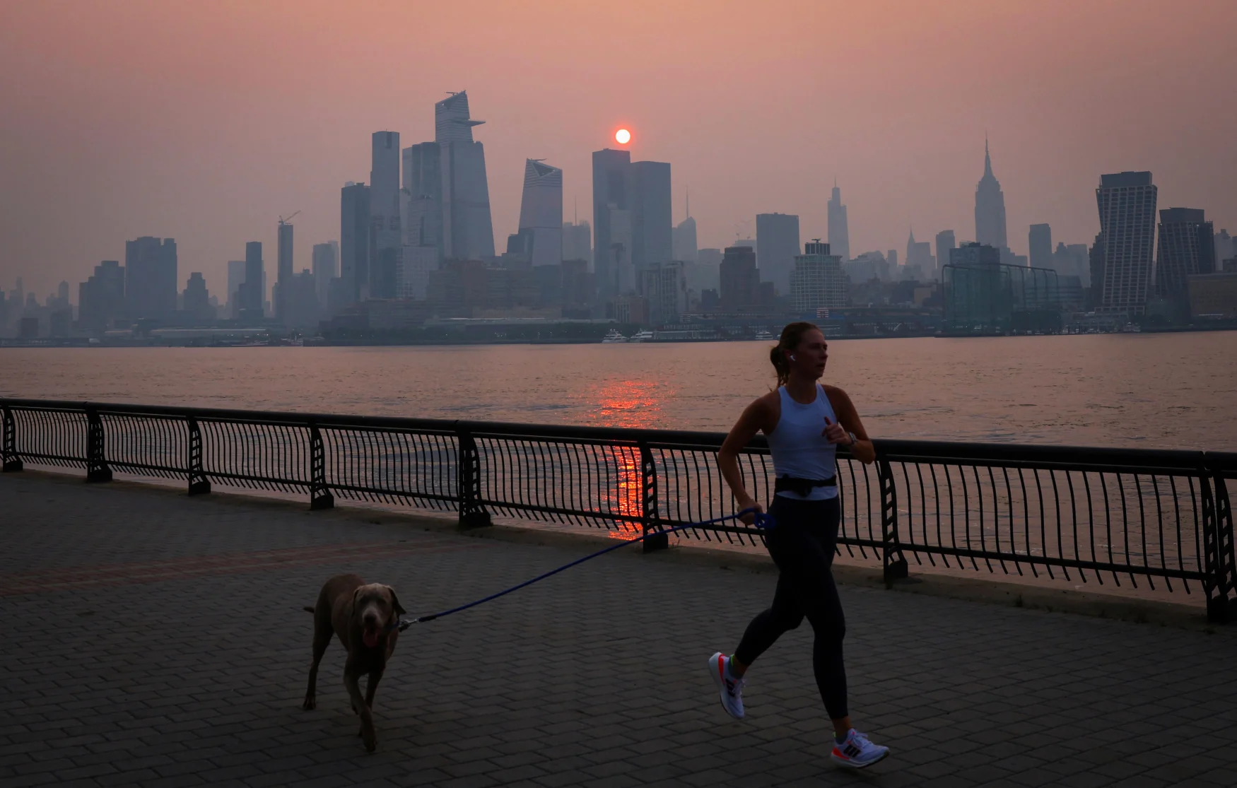 A woman jogs with a dog along the Hudson River shortly after sunrise, as haze and smoke caused by wildfires in Canada hang over the Manhattan skyline, in New York City, New York, U.S., June 7, 2023. REUTERS/Mike Segar