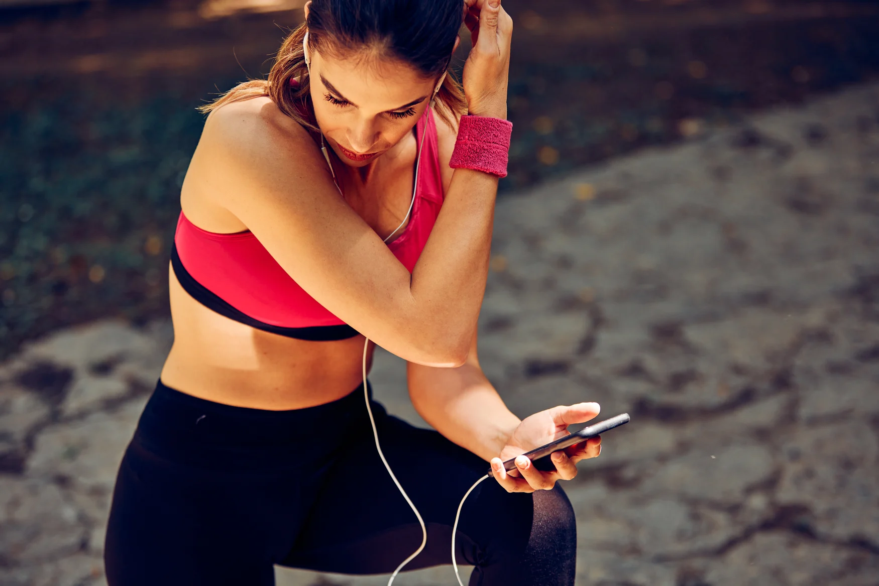 Smiling sportswoman crouching, taking a break, and using phone after running in nature.