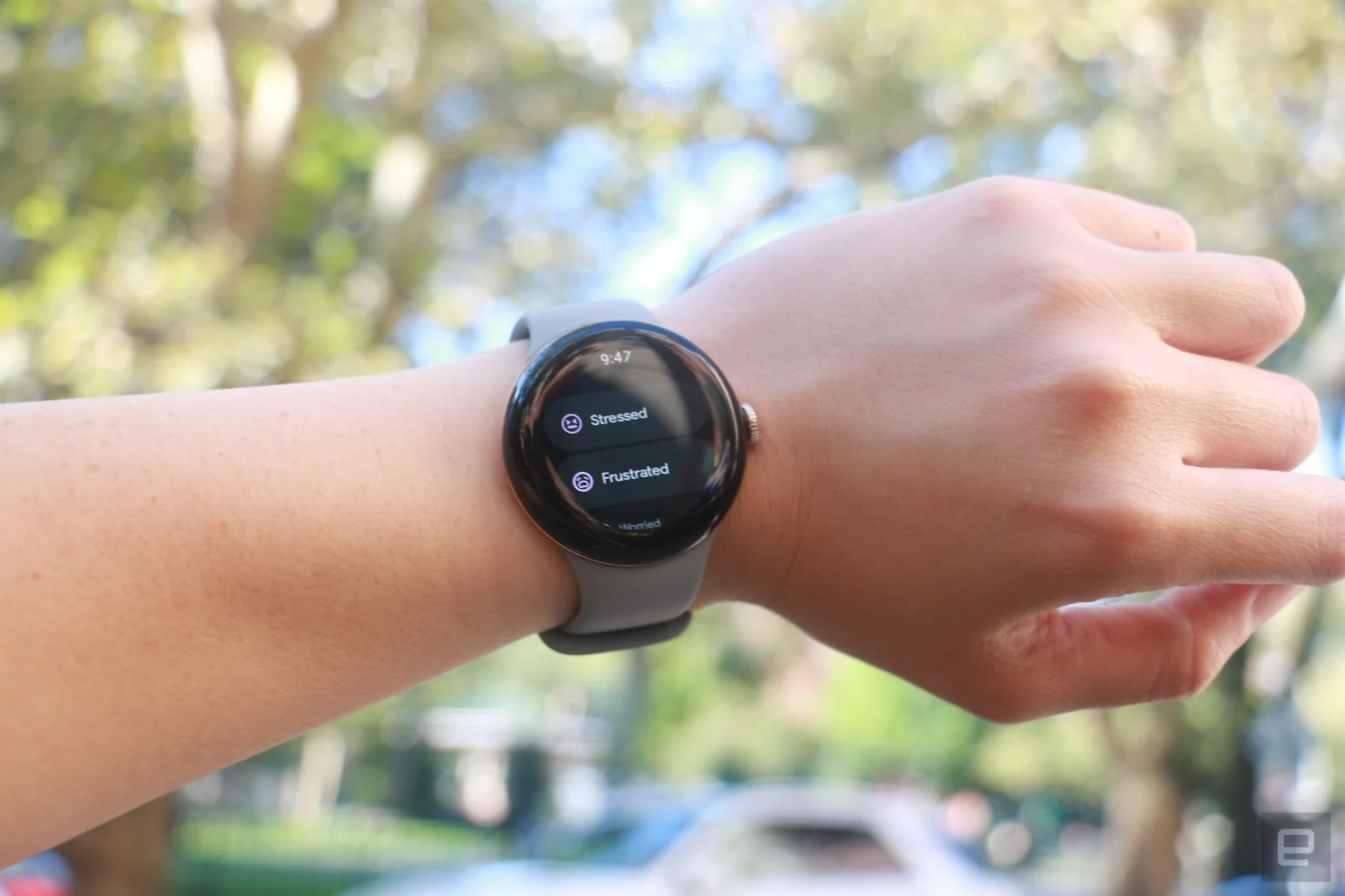 The Pixel Watch 2 on a person's wrist held up in mid-air, with some of the surrounding scenery reflecting off the screen. On the display are options for logging the user's mood, including the words 